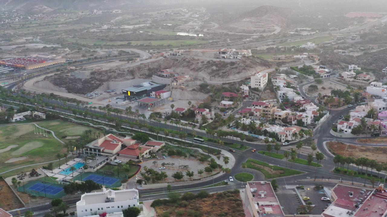 Aerial view over San Jose del Cabo showing curving roads, residential buildings, golf-course grass, tennis courts, dry hills, and no visible people.