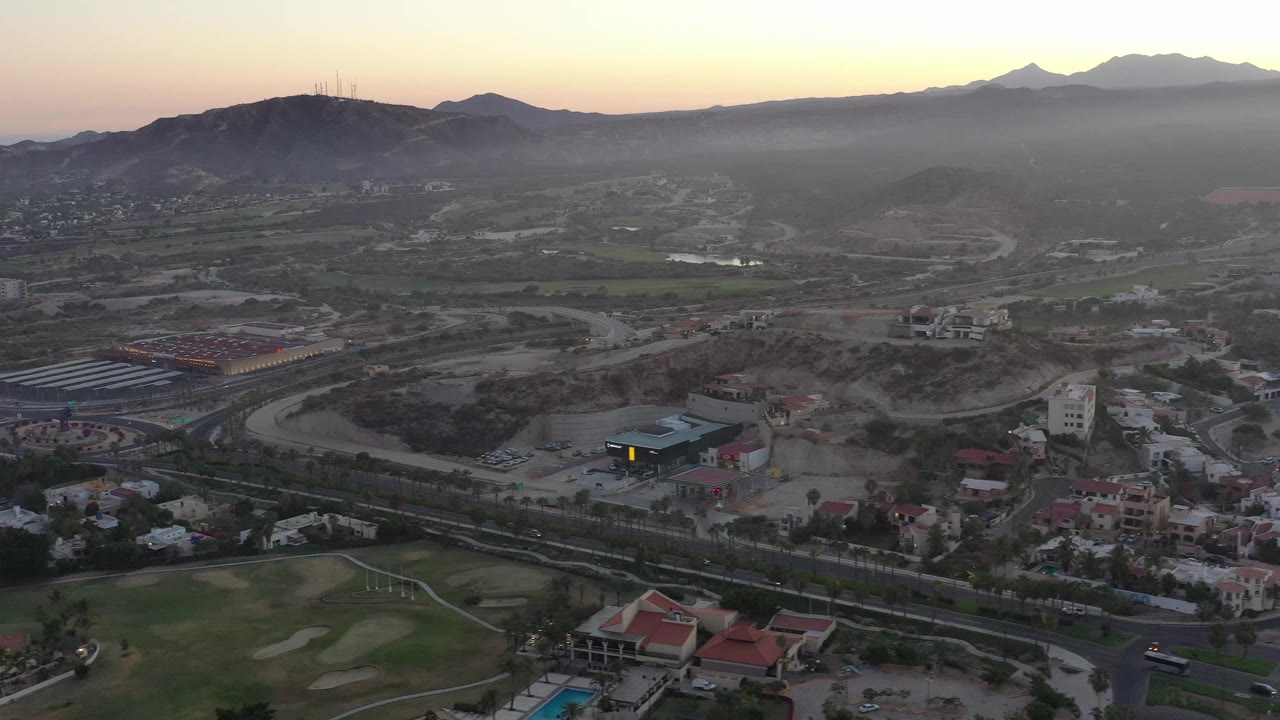 San Jose del Cabo aerial frame with residential rooftops in the lower edge, a curving road network through the midground, hazy mountains beyond, and no visible people.