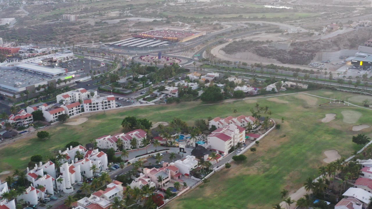 Opening aerial view in San Jose del Cabo showing white villas with red roofs beside green golf fairways, roads, and larger buildings in the distance, with no visible people.