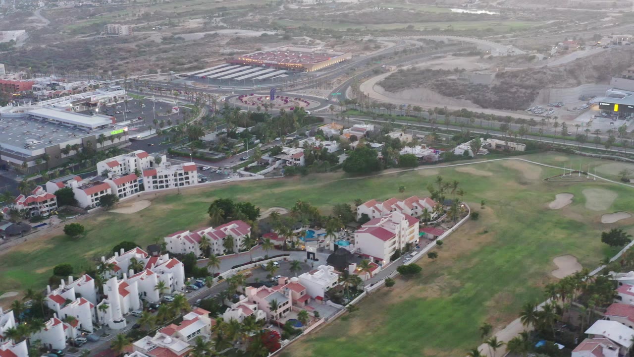 Aerial frame in San Jose del Cabo with red-roof residential blocks in the foreground, golf-course greens across the center, and town roads and commercial structures beyond, with no visible people.