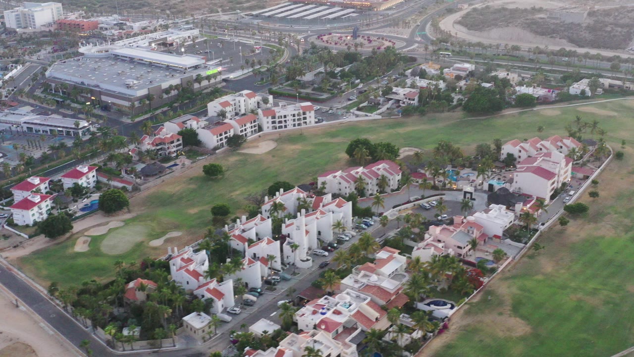 A high aerial view over San Jose del Cabo shows green fairways, dry sand areas, white residential buildings with red roofs, roads, parked cars, and modern commercial blocks, with no visible people.