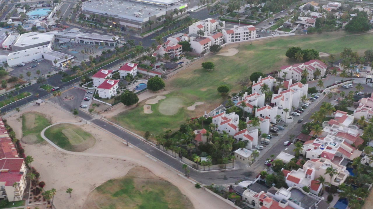 Opening high aerial frame over San Jose del Cabo showing red-roofed homes, curved roads, golf course sand traps, pools, palms, and no visible people.