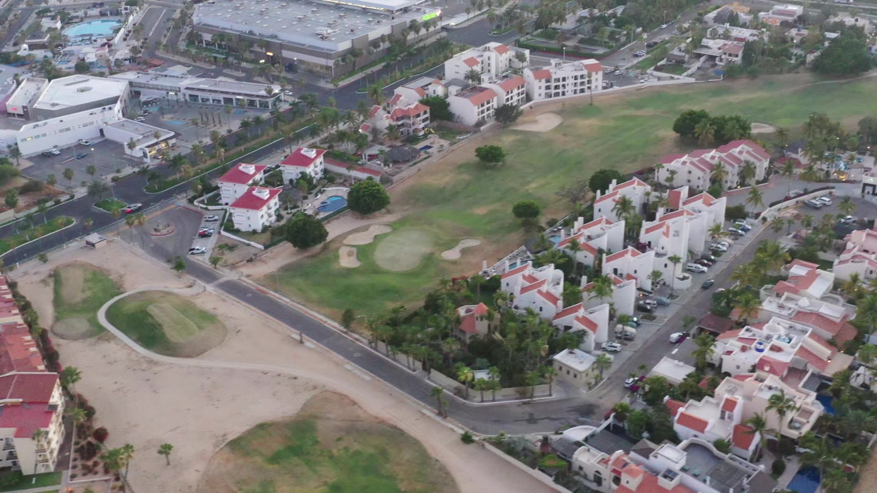 Second aerial frame in San Jose del Cabo with a broader view of the fairway, red-roofed residential buildings, roads, pools, parked cars, and no visible people.