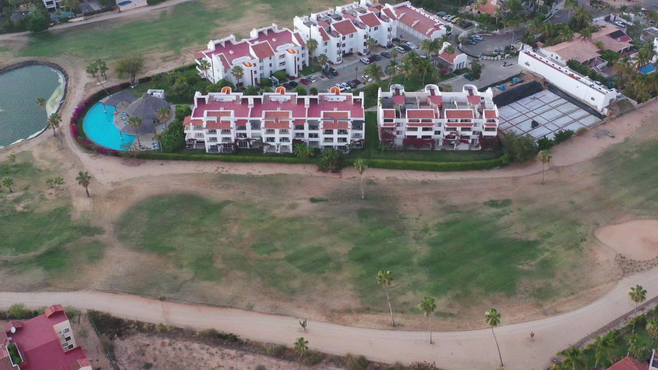 High aerial frame over San Jose del Cabo residences with red roofs, a bright blue pool at left, open grass below, and no visible people.