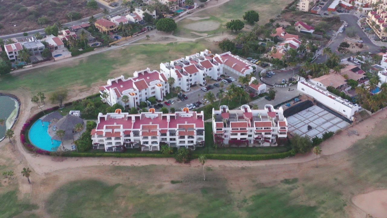 Aerial view in San Jose Del Cabo, Mexico, showing white condo buildings with red tile roofs, a bright blue pool at left, green grounds, roads, and no visible people.