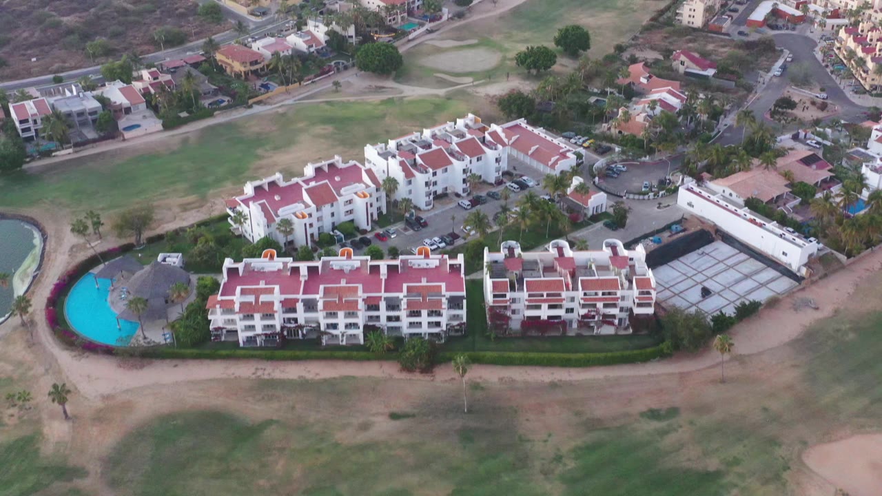 High aerial frame in San Jose Del Cabo, Mexico, with red-roofed apartment blocks centered, the turquoise pool on the left edge, neighborhood roads above, and no visible people.