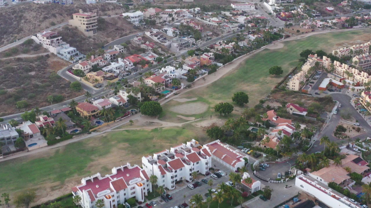 Aerial frame over a San Jose del Cabo residential area with red-roofed homes in the foreground, curving roads, dry hillside terrain, green open space, and no visible people.