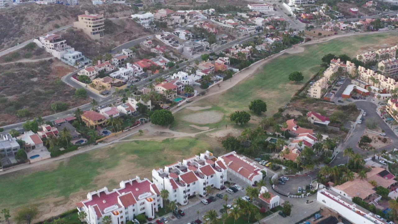 Aerial view in San Jose del Cabo showing white residences, red tile roofs, a road network, a green fairway-like open space, dry hills, distant buildings, and no visible people.