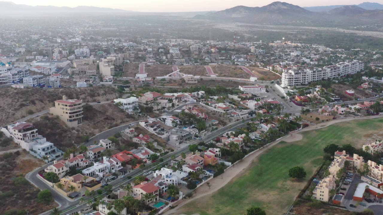 High view across San Jose del Cabo with a curved road and neighborhood roofs in the lower frame, a broad green fairway to the right, town buildings beyond, and no visible people.