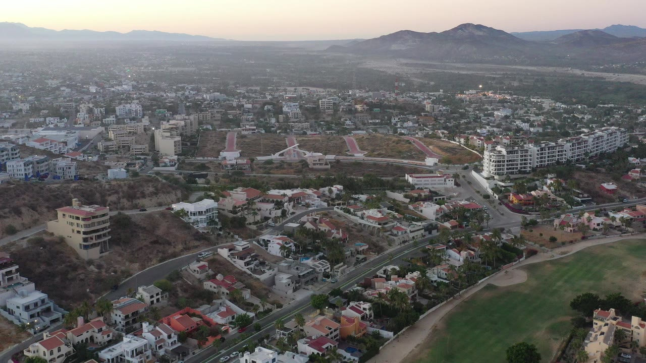 Early aerial frame over San Jose del Cabo, Mexico, showing residential rooftops, curved roads, a green open area, distant mountains, hazy evening light, and no visible people.