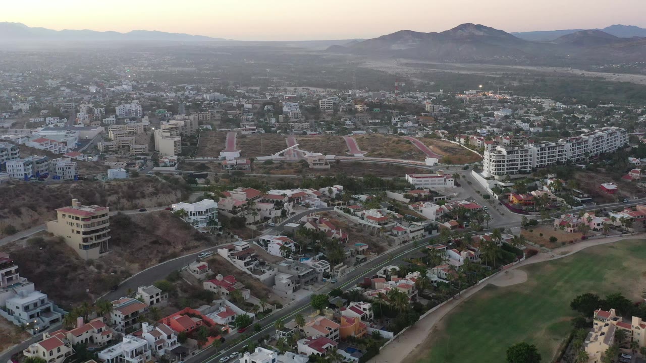 Aerial view over San Jose del Cabo, Mexico, with a denser cluster of low-rise buildings in the center, homes below, mountains on the horizon, and no visible people.