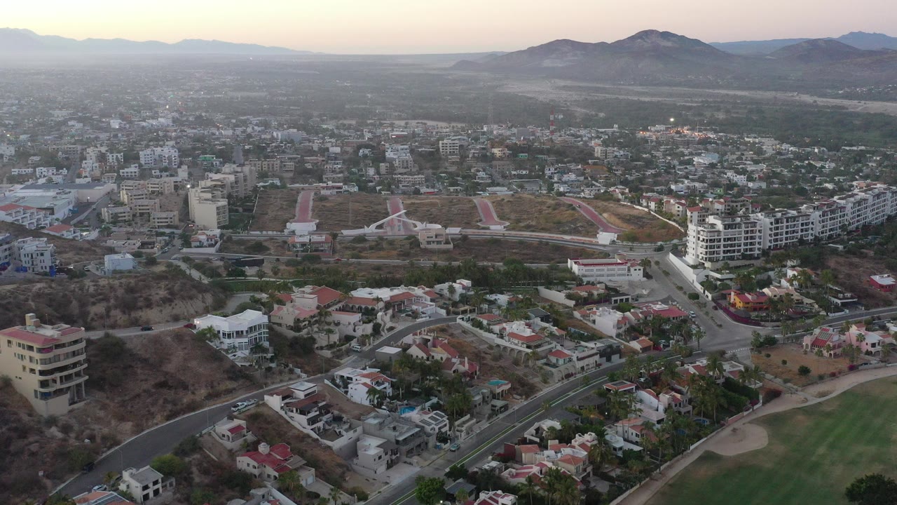 Wide aerial view of San Jose del Cabo, Mexico with red-roof homes, apartment buildings, dry open lots, roads, distant mountains, hazy sky, and no visible people.