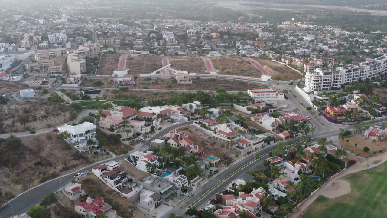 High drone view over San Jose del Cabo houses, curving roads, a green fairway edge, dry hillside lots, and distant buildings, with no visible people.
