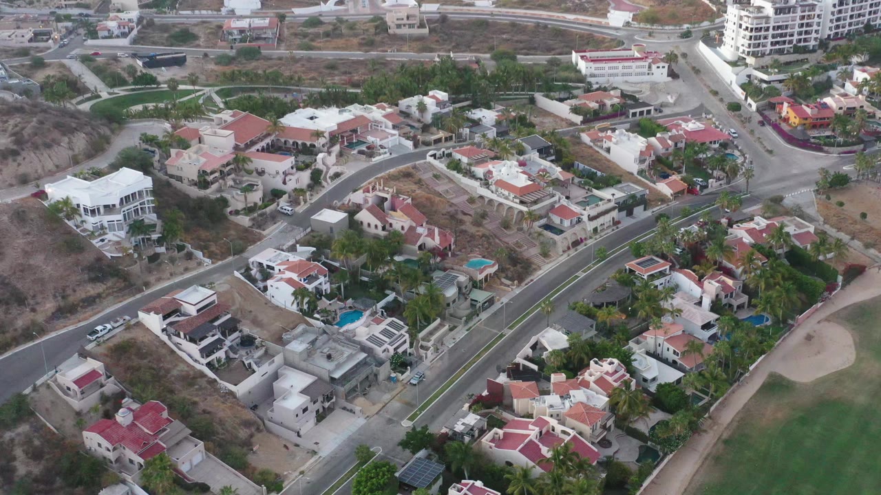 High aerial frame over San Jose del Cabo homes, showing white and terracotta roofs, palms, a sloping road, dry open parcels, and no visible people.