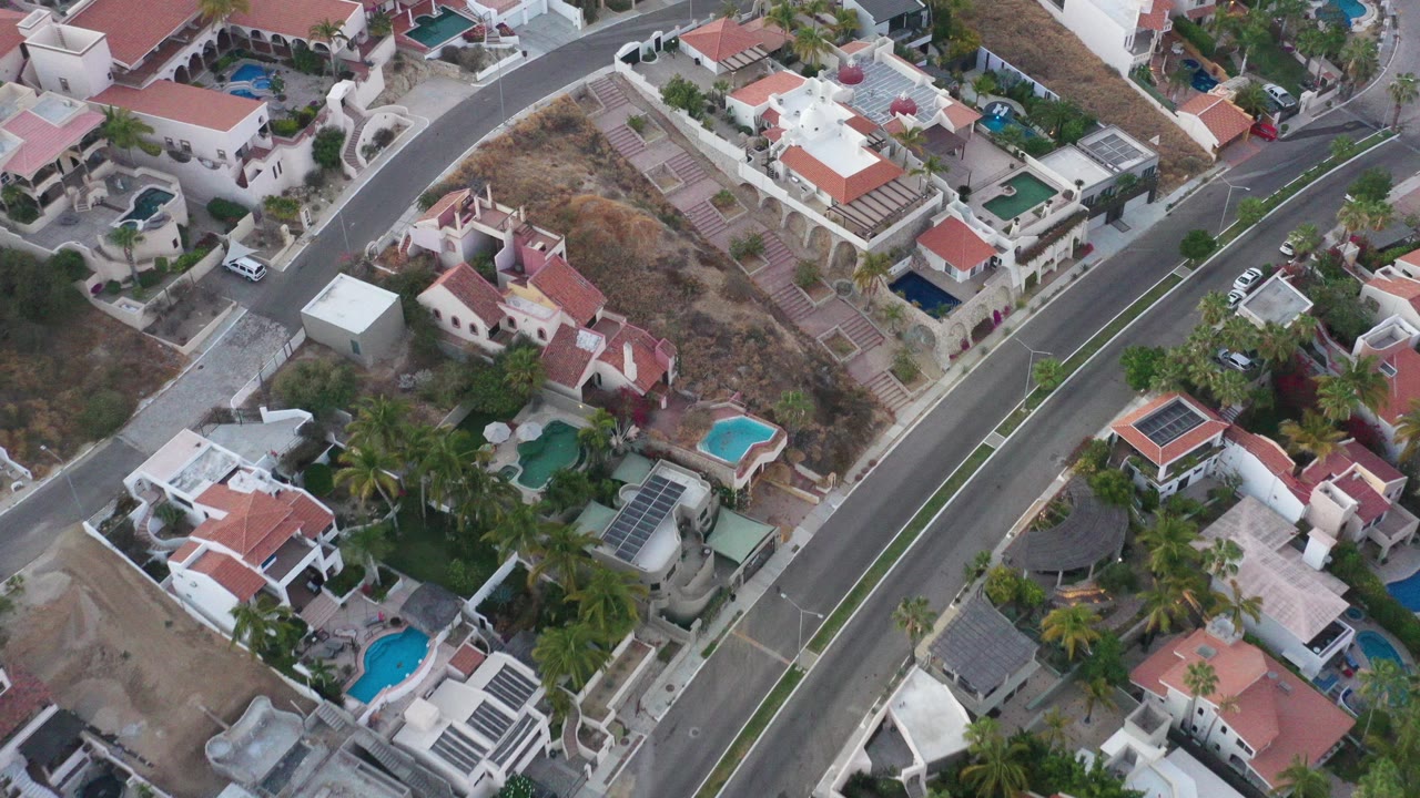 Aerial frame over San Jose del Cabo homes with red tile roofs, turquoise pools, palm trees, a diagonal paved road, and no visible people.