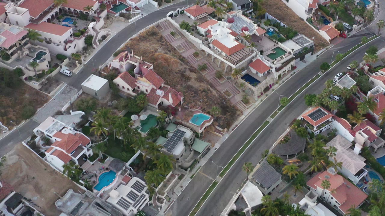 Aerial frame over San Jose del Cabo showing walled residential properties, several pools, parked vehicles, dry ground, and no visible people.