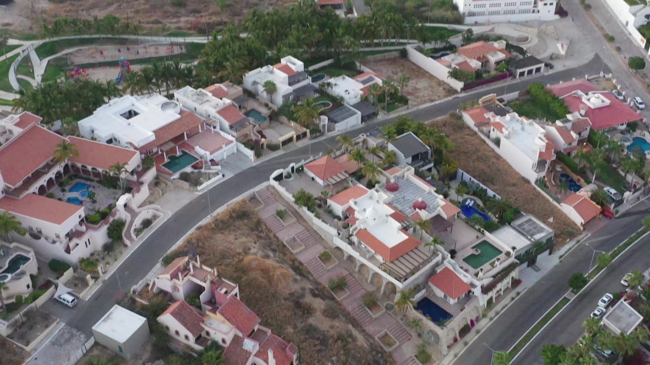 High aerial view in San Jose del Cabo showing a curved residential road, white villas, red roofs, palms, dry ground, parked cars, and no visible people.