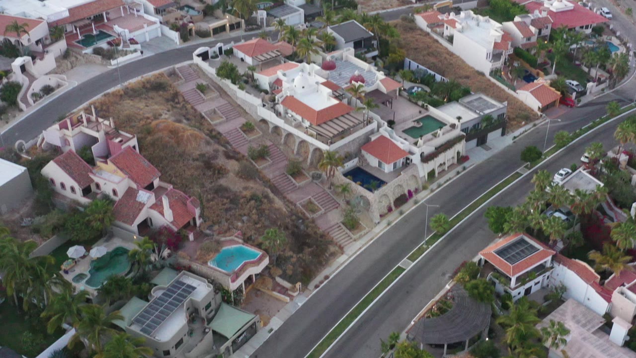 Aerial view in San Jose Del Cabo centered on white villas, a divided road, turquoise pools, terraced dry ground, and no visible people.