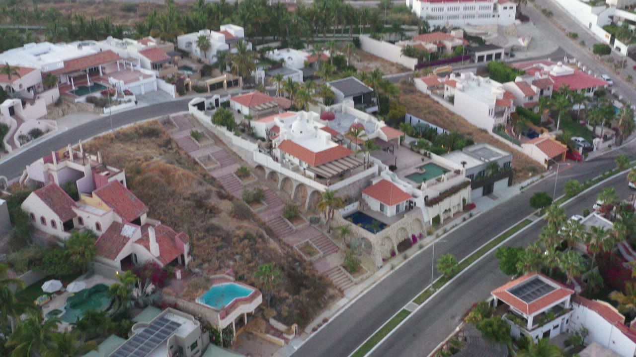 Aerial view of white villas, red tile roofs, curved road, palms, and swimming pools.