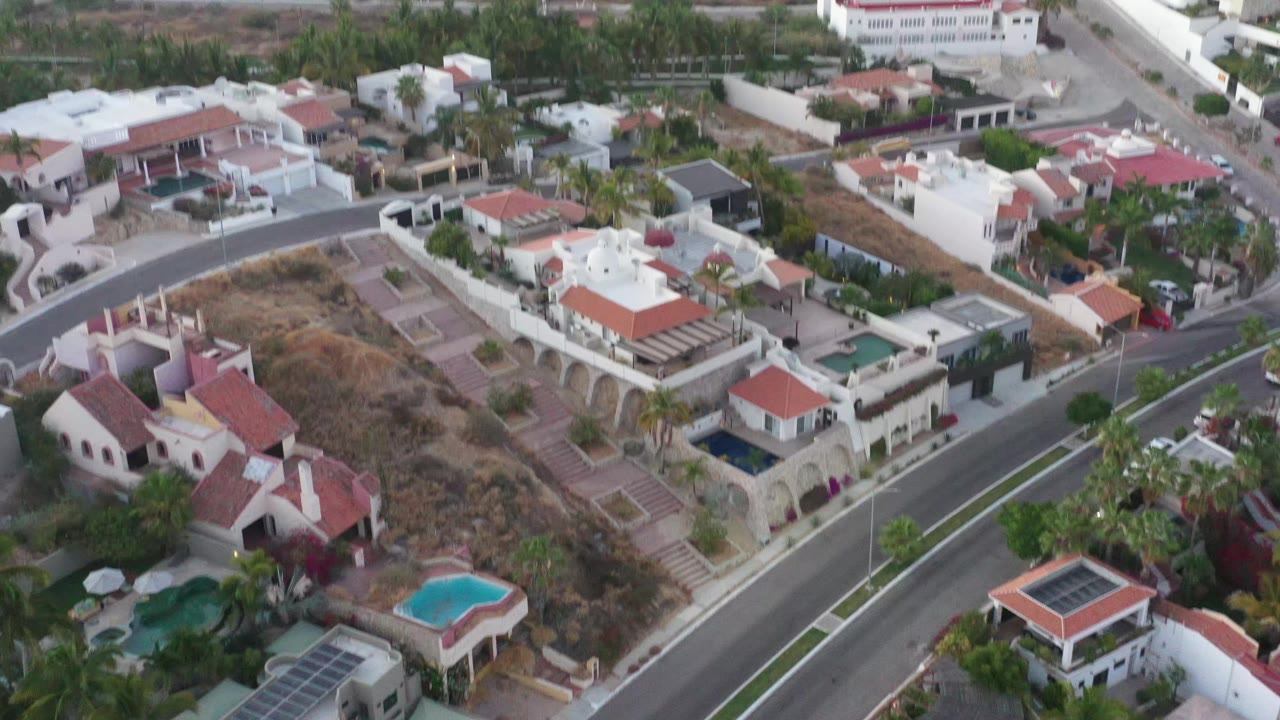 Aerial frame in San Jose del Cabo showing villas and garden walls beside dry open parcels, with palms and small pools visible and no visible people.
