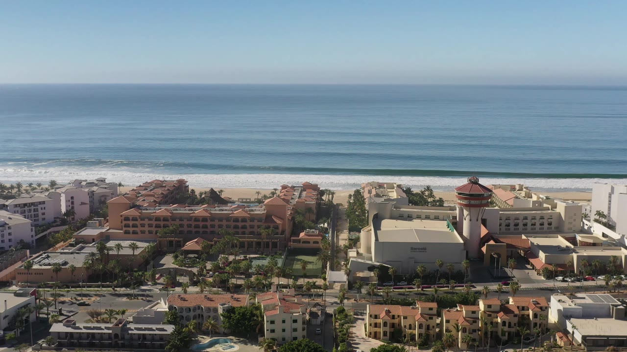 Aerial view in Los Cabos, Mexico, with the resort complex shifted slightly across frame, white surf bands behind it, and no visible people.