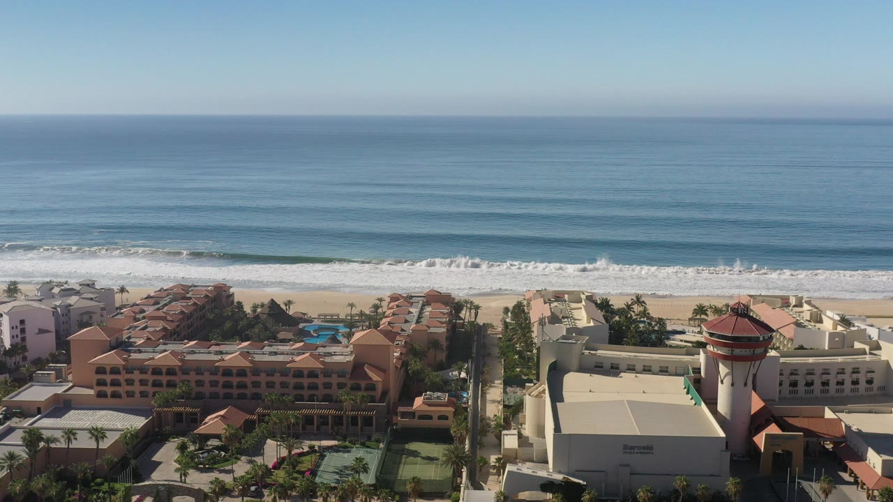 Aerial view in Los Cabos, Mexico showing resort roofs, palms, a sandy beach, rolling surf, blue ocean, and no visible people at the start of the clip.