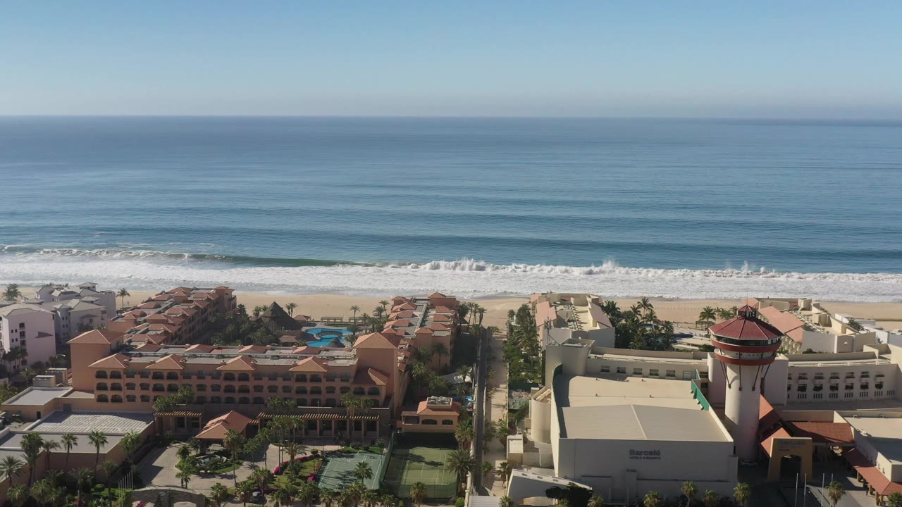 Aerial frame in Los Cabos, Mexico with more of the central resort roofs and pool visible below the beach, surf bands, open water, and no visible people.