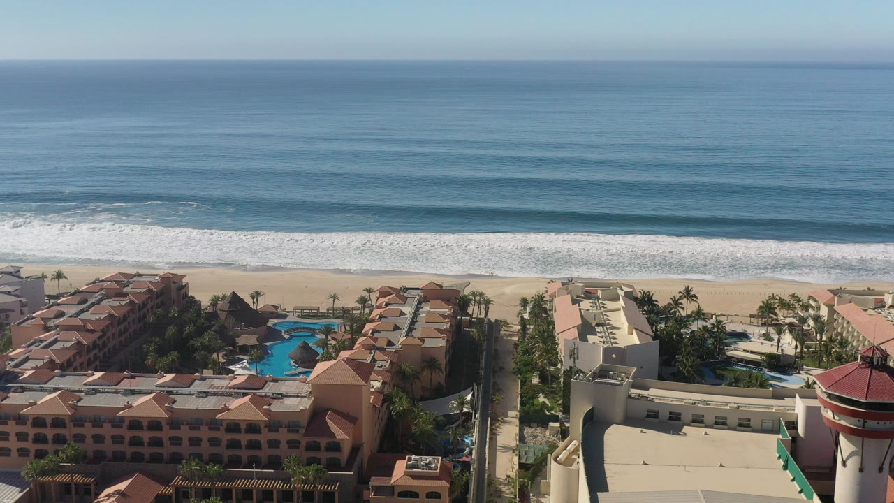 Aerial frame over Los Cabos, Mexico showing red-roofed beachfront buildings, blue swimming pools, palms, sand, white surf, open ocean, and no visible people.
