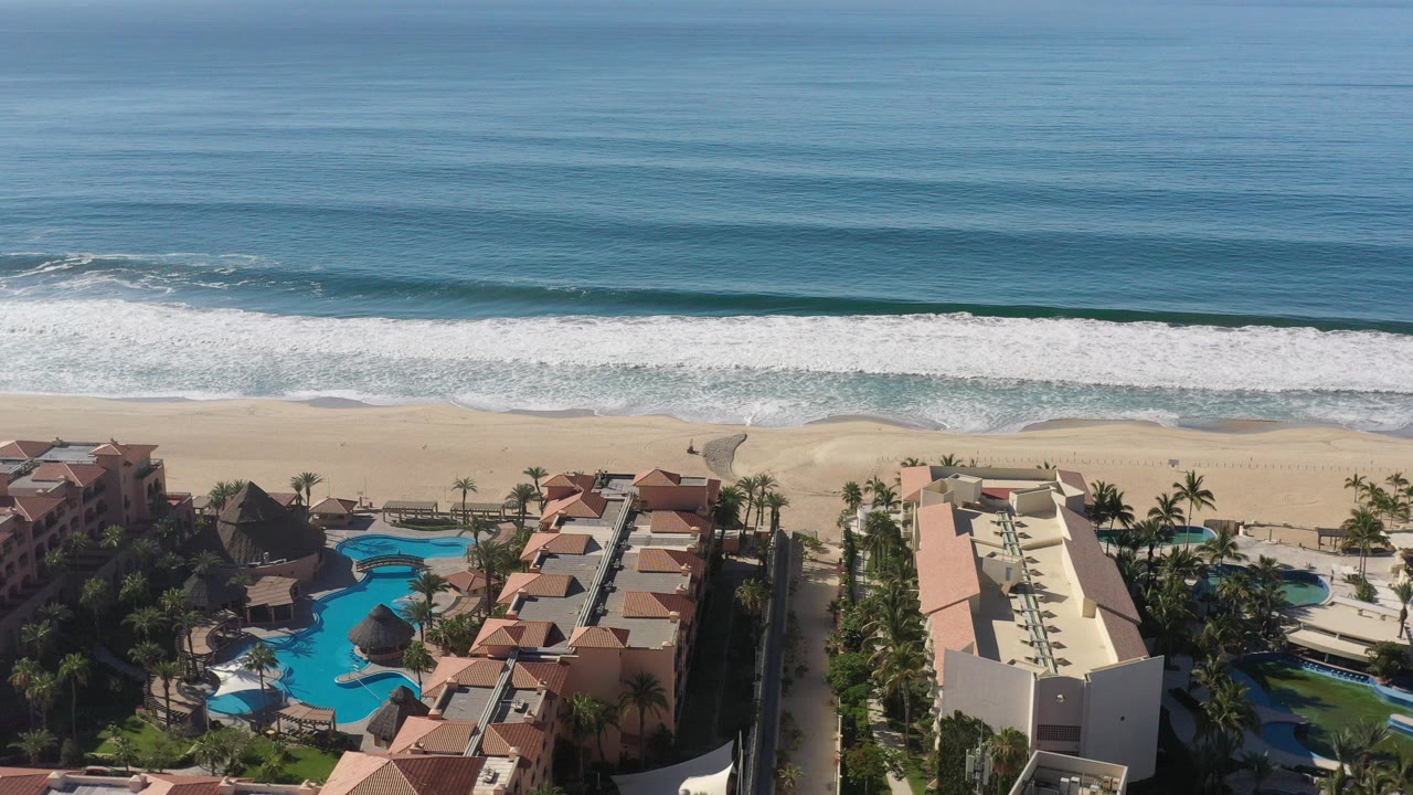 High aerial view over a Los Cabos beach with deep blue ocean, a white surf line, sand, resort buildings, pools, palm trees, and no visible people.