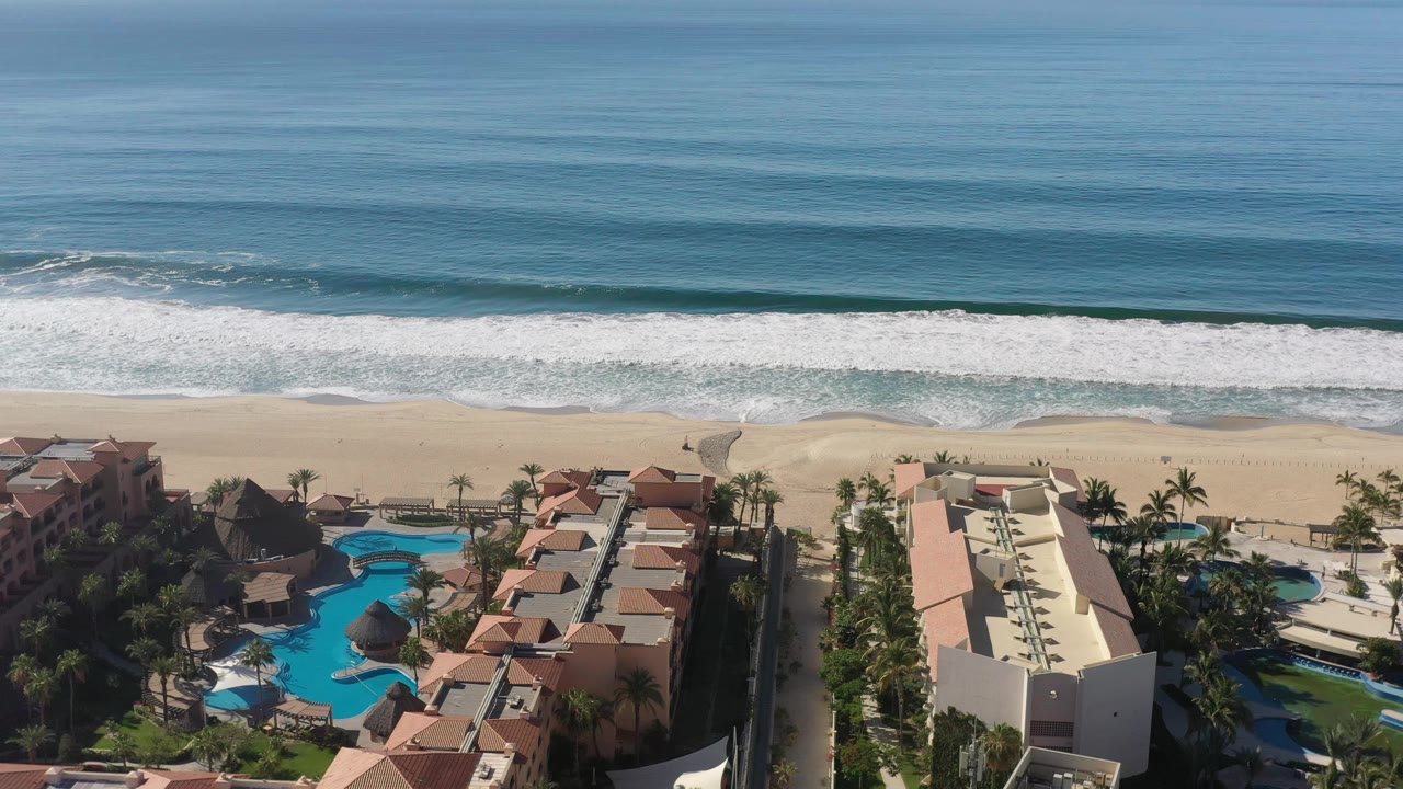 Elevated Los Cabos coastline frame showing rolling surf, a wide sandy beach, hotel blocks, bright blue pools, palms, and no visible people.