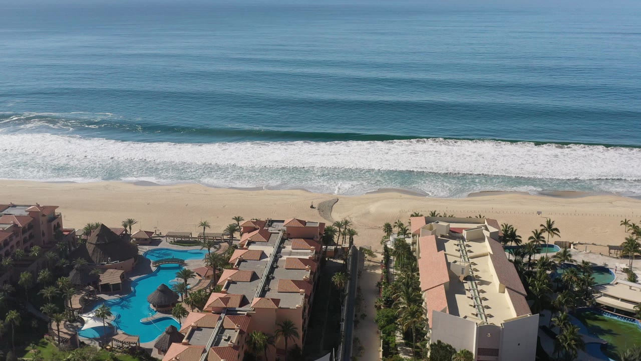 Top-side aerial view of ocean, surf, sand, hotels, pools, and palms.