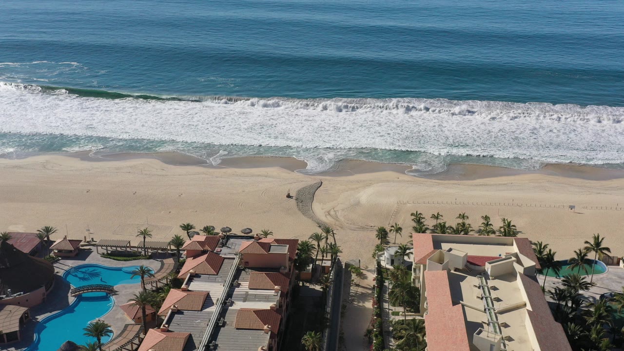 Aerial view of surf, sandy beach, resort pools, rooftops, and palm trees.