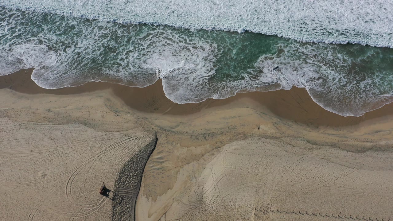 Overhead view in Los Cabos, Mexico, with foamy surf across the top of frame, green-blue water at the break, tan sand below, and no visible people.