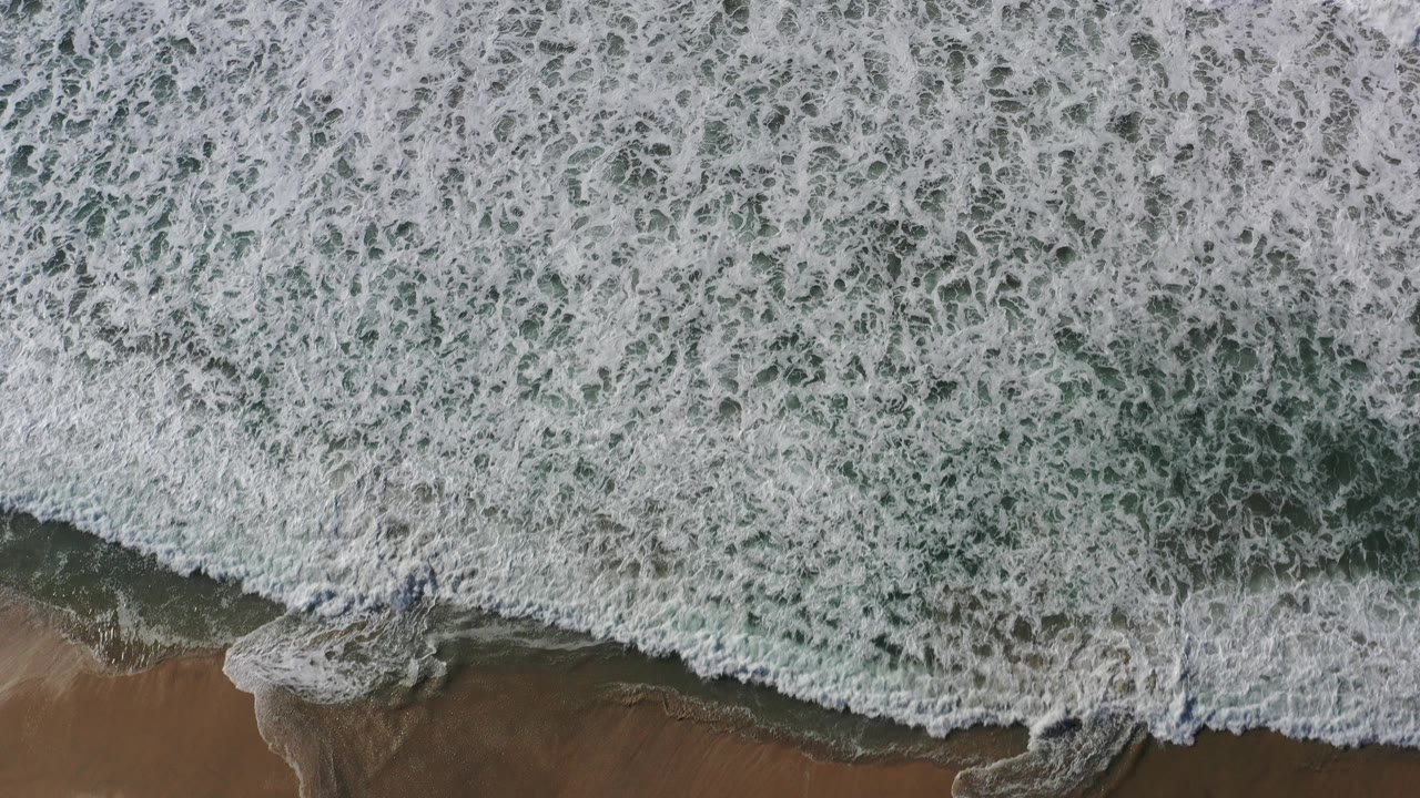 Overhead drone view of white surf covering green-blue water beside a thin band of sand in Los Cabos, Mexico, with no visible people.