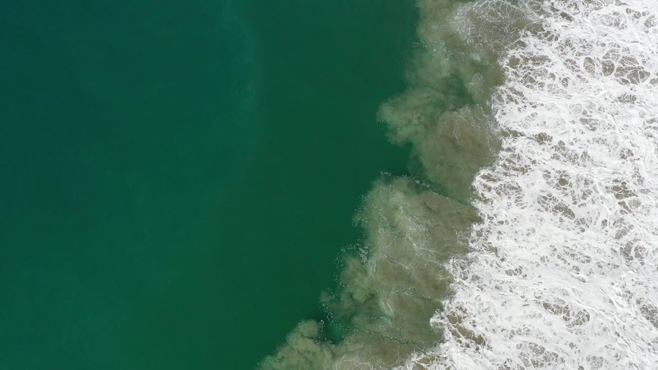 Overhead view in Los Cabos, Mexico, with dark green ocean filling the left side and white shore break entering from the right, no visible people.