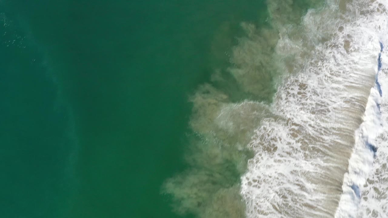 Aerial Los Cabos surf frame with open green water on the left and a bright white wave edge spreading over cloudy tan sand, with no visible people.