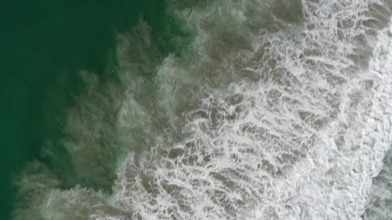 Overhead view of green ocean water in Los Cabos, Mexico, with dense white foam pushing across the right side and no visible people.