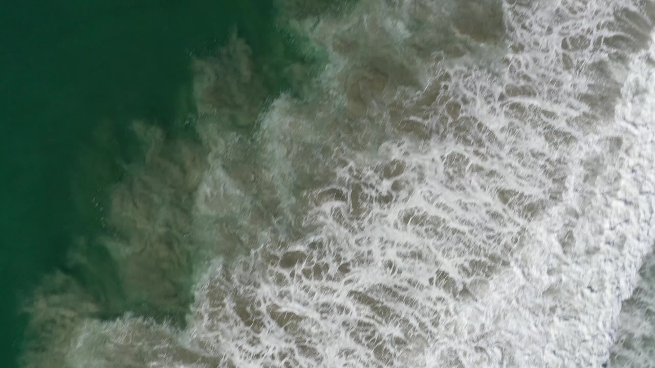 Overhead surf frame from Los Cabos, Mexico, showing whitecaps spreading through green water with a darker open patch on the left and no visible people.
