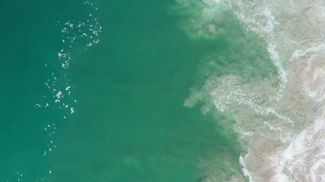 Overhead view in Los Cabos, Mexico with turquoise ocean on the left, pale rocky shoreline on the right, bright water sparkle near the upper left, and no visible people.