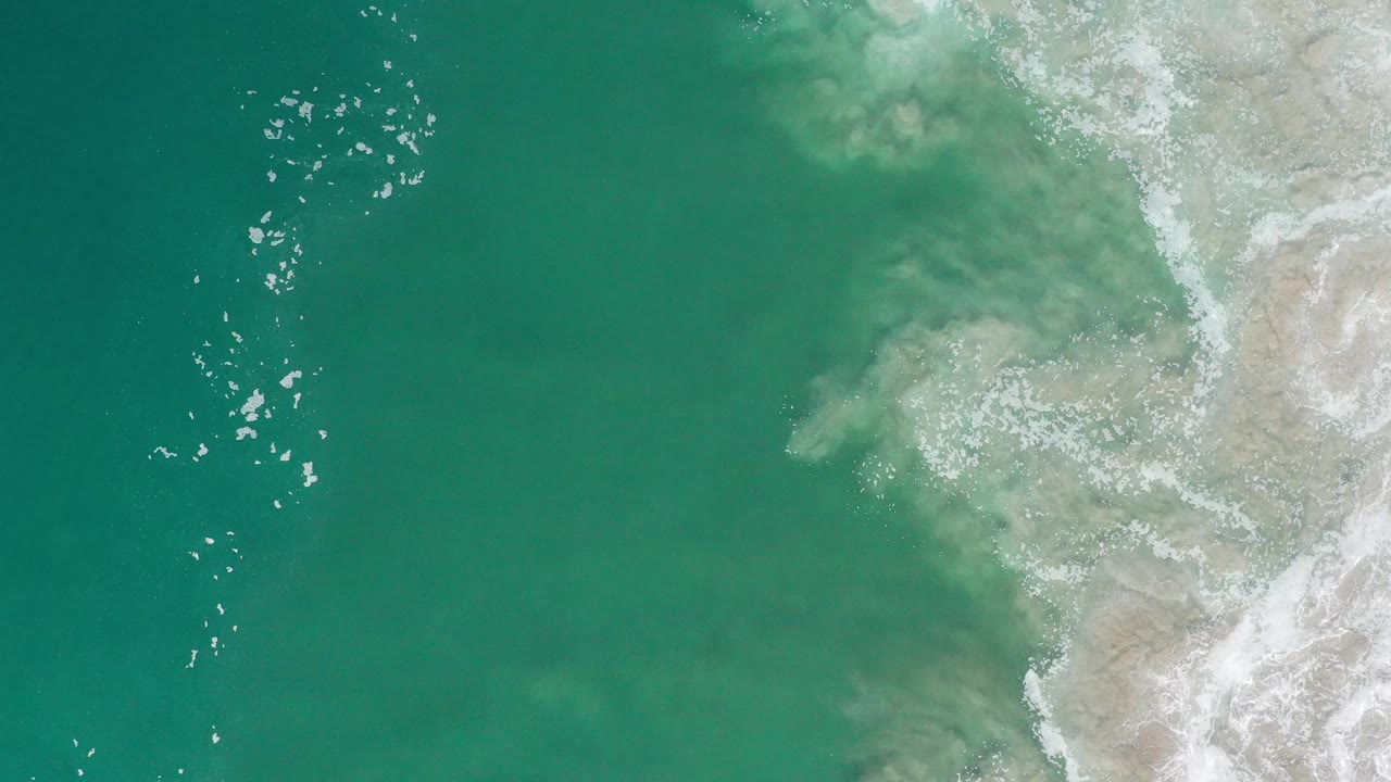 High overhead Los Cabos frame with open green-blue water, a pale coast along the right side, scattered white foam, and no visible people.