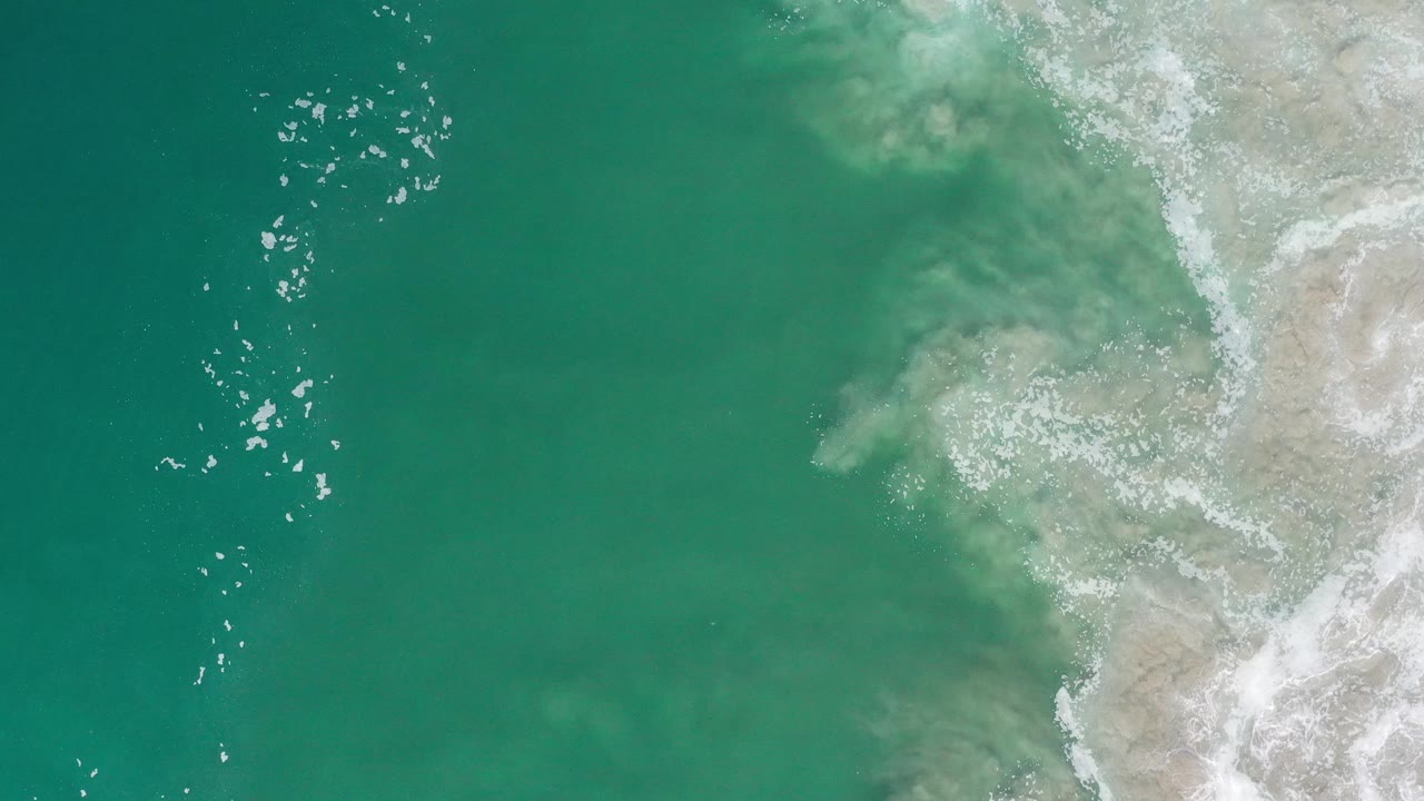 Turquoise ocean and irregular white surf along a rocky shoreline from above.