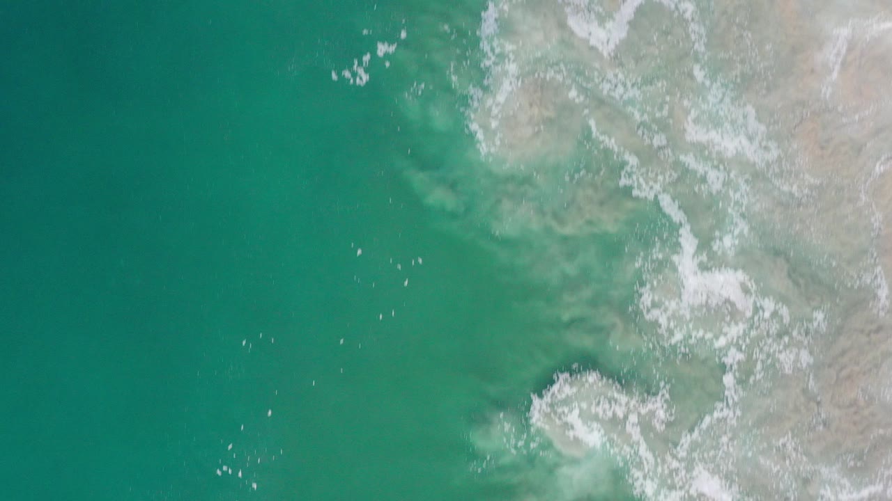 Overhead turquoise ocean beside pale rocky shoreline and white surf.