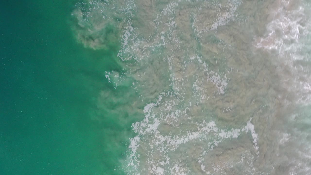 High overhead view of turquoise water meeting a pale Los Cabos shoreline with scattered white foam and no visible people.