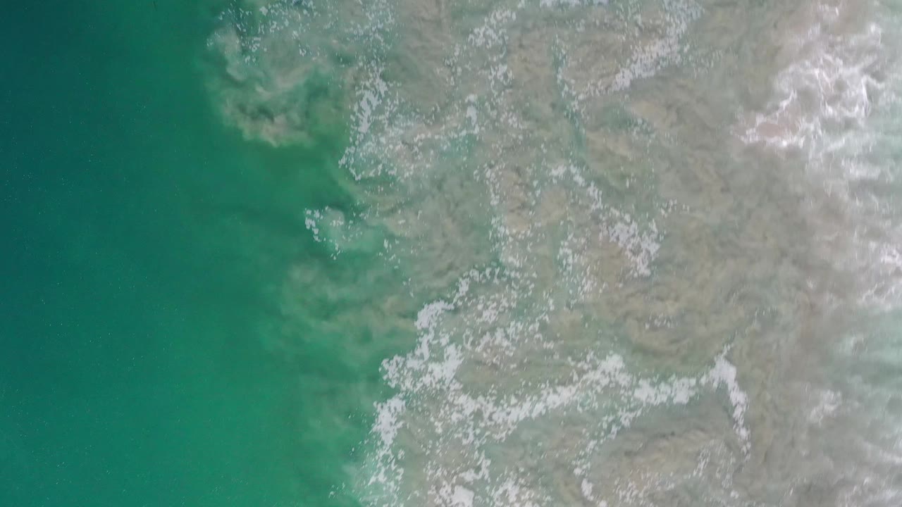 Overhead Los Cabos coastal frame with blue-green water on the left, pale shore texture on the right, and no visible people.