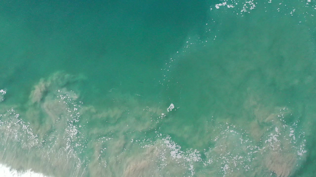 Overhead view of teal ocean water in Los Cabos, Mexico, with a pale submerged sand plume, scattered white foam, and no visible people.