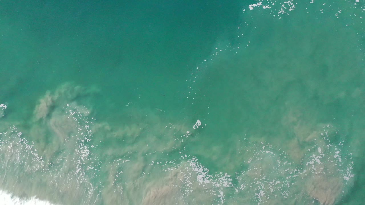 Aerial frame over Los Cabos ocean water showing turquoise surface color, a broad tan sediment swirl, bright foam flecks, and no visible people.