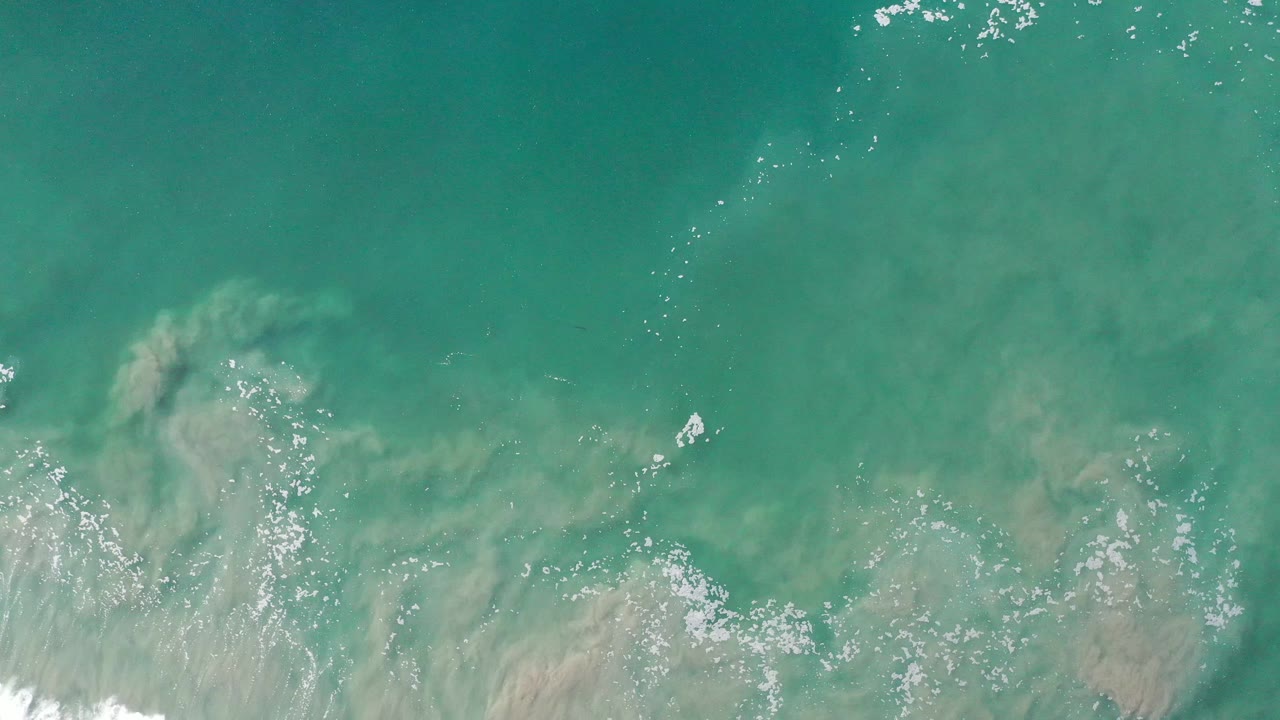 Overhead ocean surface with sand cloud and white foam patches.