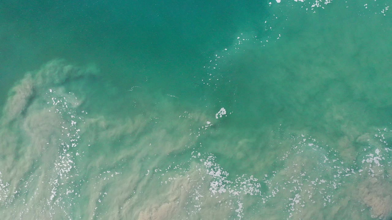 Overhead view of blue-green coastal water in Los Cabos, Mexico, with pale sandy swirls under the surface, small foam patches, sun glints, and no visible people.