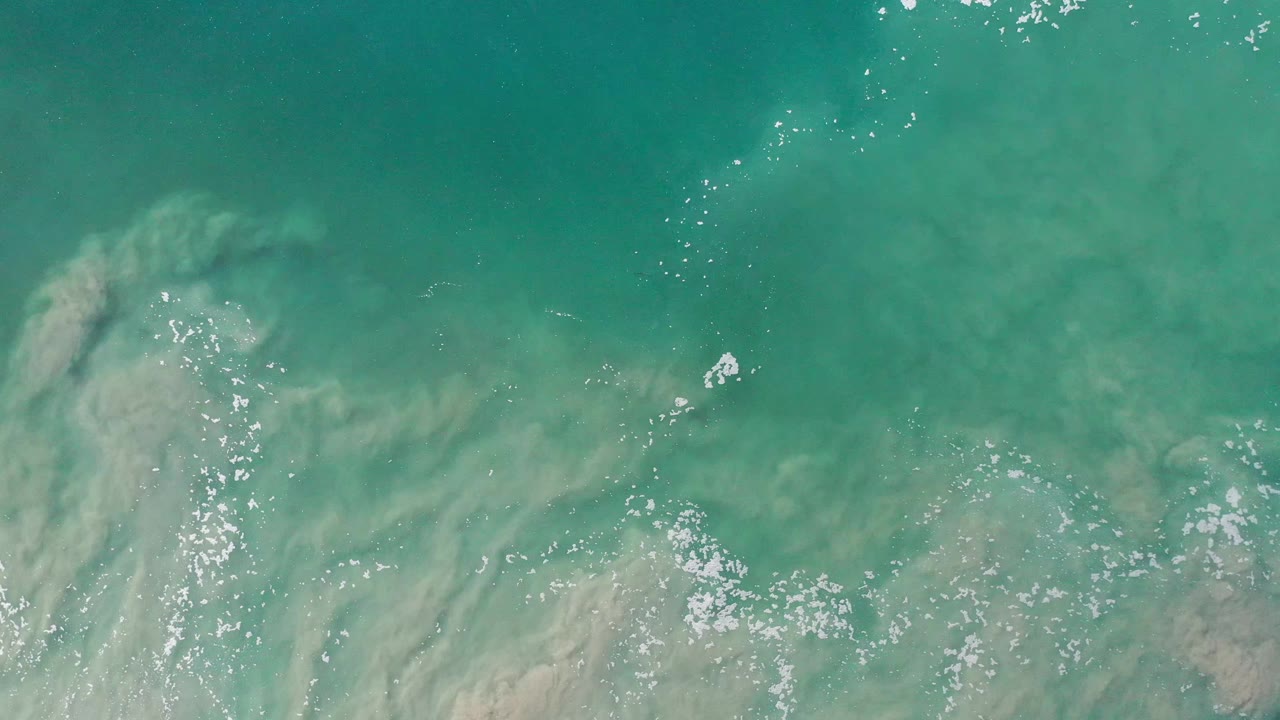 A top-down Los Cabos water frame shows a curved pale sand plume below the turquoise surface, bright white foam specks, ripples, and no visible people.