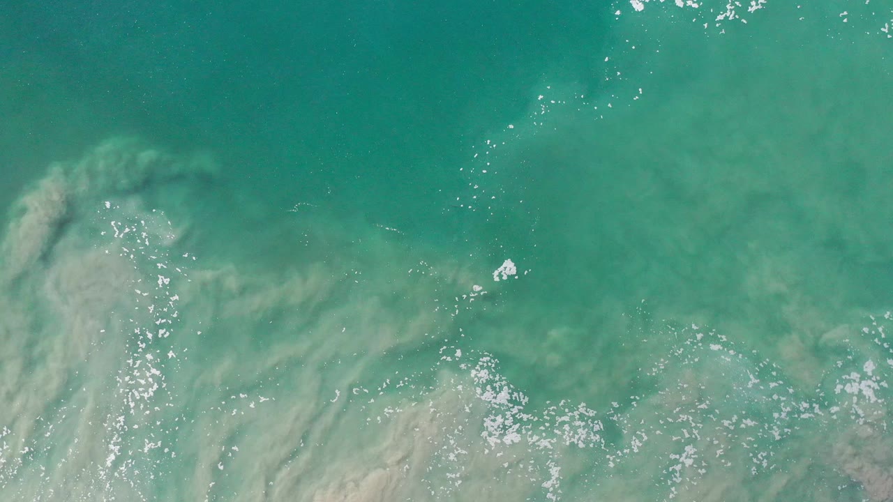 Turquoise coastal water with underwater sand haze and scattered sparkling foam.